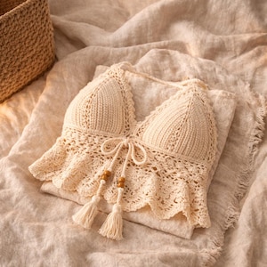 Peut inclure: Haut en crochet de couleur cr&egrave;me avec un col halter et un ourlet &agrave; volants. Le haut pr&eacute;sente une fermeture &agrave; nouer avec des perles en bois et des pompons. Le v&ecirc;tement fait main est pr&eacute;sent&eacute; sur un tissu aux tons neutres, mettant en valeur ses d&eacute;tails et sa texture.