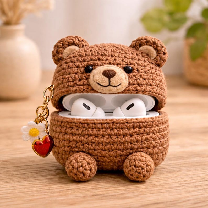 May include: A brown crochet bear case for wireless earbuds featuring a smiling bear face with round ears and two small feet at the base. A gold-toned chain is attached to the side, adorned with a small white daisy charm and a shiny red heart charm.