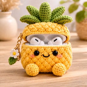 May include: A yellow crochet pineapple case for wireless earbuds featuring a green leafy top, a smiling face with black eyes and pink cheeks, and two small feet. A gold-toned chain with a white flower and green leaf charm is attached to the side.