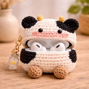 May include: A crochet cow-shaped case for wireless earbuds, featuring a black and cream colour scheme, a pink snout, two small yellow horns, and two brown feet. A gold-toned chain with a small clear jar charm containing a white flower is attached.