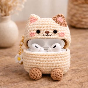 May include: A cream-coloured crochet case for wireless earbuds shaped like a cat with brown ear accents, pink cheeks, and two small brown feet at the base. The case features a gold-toned metal keychain with a small white flower charm attached.