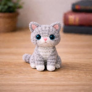 May include: A handmade crochet cat figurine in shades of gray and white. The cat has large, blue eyes, pink inner ears, and a pink nose. The cat is sitting on a wooden surface.
