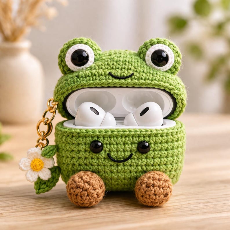 May include: A green crocheted frog-shaped case for wireless earbuds. The case features large black eyes, a stitched smile, and two brown feet at the base. A gold-toned metal keychain with a small white and yellow crocheted flower is attached.