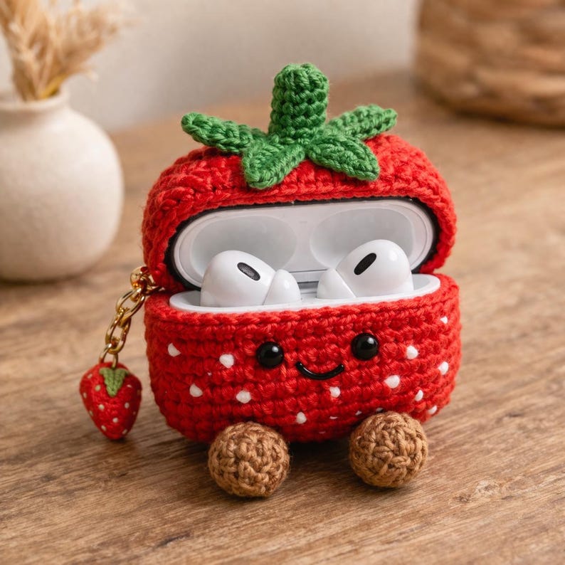 May include: A red crocheted strawberry-shaped case for wireless earbuds. The case features a green stem top, white embroidered dots, two black bead eyes, a stitched smile, and two small brown feet. A miniature strawberry charm hangs from a gold chain.