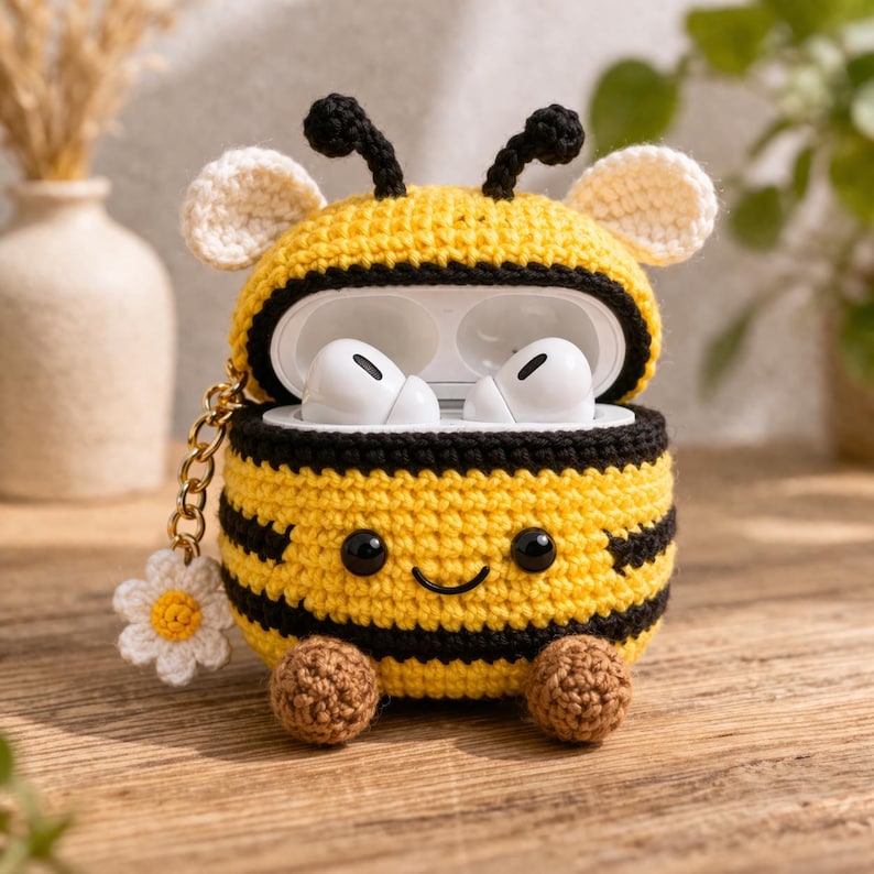 May include: A crochet bee-shaped case for wireless earbuds, featuring yellow and black stripes, two black antennae, white wings, and two brown feet. The case has a smiling face with black eyes and includes a gold chain with a white flower charm.