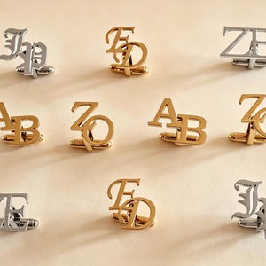 Puede incluir: Una colección de gemelos con monogramas personalizados en tonos dorados y plateados. Cada pieza presenta dos letras entrelazadas en varias fuentes, incluyendo estilos serif, sans-serif y góticos, dispuestos en tres filas sobre una superficie clara.