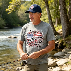 Puede incluir: Una camiseta gris de manga corta con un estampado gráfico de una bandera estadounidense, una caña de pescar y un pez, combinada con una gorra de béisbol azul marino que tiene un parche circular con un diseño de pesca y bandera.