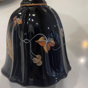 May include: A dark blue bell-shaped glass with a flared base and a long stem. The bell is decorated with orange and white floral designs. The glass has a glossy finish and is held by a hand.