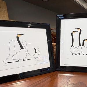 May include: Two framed art prints featuring minimalist black and white line drawings of geese with yellow accents on their heads. The left print shows two geese, while the right print displays three geese. Both prints are encased in glossy black frames.