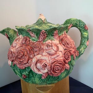 Puede incluir: Una tetera de cerámica con un relieve tridimensional de rosas rosas y rojas con hojas verdes. El asa y la tapa están esculpidas para parecer tallos con hojas, y toda la superficie está cubierta con texturas detalladas de flores y follaje.