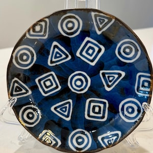 May include: A circular ceramic plate featuring a dark blue glaze with a pattern of white geometric shapes, including circles, squares, and triangles. The plate has a brown rim and is displayed on a clear stand.