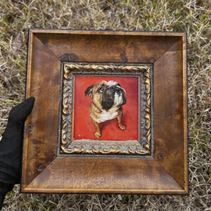 Puede incluir: Pintura enmarcada de un bulldog sobre fondo rojo. El perro es marrón, blanco y negro, con una expresión concentrada. El marco es de madera oscura con detalles dorados ornamentados en el marco interior.