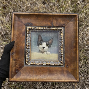 Puede incluir: Pintura enmarcada de un gato blanco y negro con ojos verdes. La obra de arte está en un marco cuadrado, ornamentado, de madera oscura con detalles dorados. El gato se asoma por encima de una superficie, creando un retrato encantador.