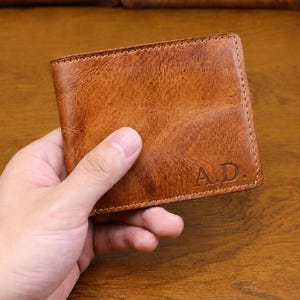 Puede incluir: Cartera de cuero marrón con las iniciales "A.D." grabadas en la esquina inferior derecha. La cartera se sostiene en una mano, mostrando su superficie texturizada y bordes cosidos. La cartera parece nueva.