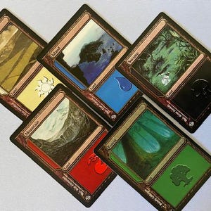 以下が含まれることがあります： 扇状に並べられた5枚のMagic: The Gatheringの土地カード。カードはPlains、Island、Swamp、Mountain、Forestです。各カードには風景のイラスト、色付きのマナシンボル、そして土地タイプの名前がはっきりと印刷されています。