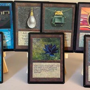 May include: A collection of eight Magic: The Gathering trading cards displayed on small wooden easels. The cards feature various illustrations including a blue lotus, a purple diamond-shaped artifact, and a stone pyramid, with printed text.
