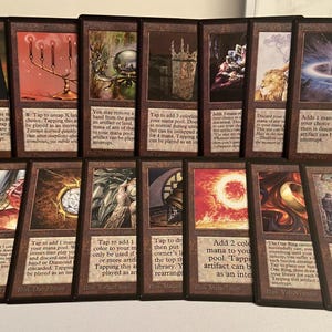 May include: A collection of fifteen rectangular trading cards arranged in two rows. Each card features a dark brown border, a unique colorful illustration in the upper section, and printed text in the lower section describing game mechanics.