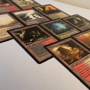 May include: A collection of Magic: The Gathering trading cards, featuring various landscape illustrations and text. The cards have a brown border and are arranged on a white surface. Some cards have text that reads "Tap to search your library".
