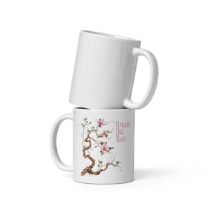 Può includere: Due tazze di ceramica bianca impilate. La tazza inferiore presenta un disegno ad acquerello di un albero in fiore con fiori rosa e il testo "Flowers Dirt Trees". La tazza superiore è bianca.