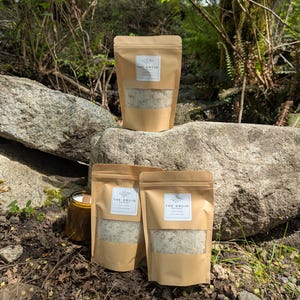 May include: Three brown paper bags with clear windows, filled with a light-coloured product, are displayed on a rock. The bags have a white label that reads "THE DRUID". A small glass jar with a gold lid is also visible. The background is a natural outdoor setting.