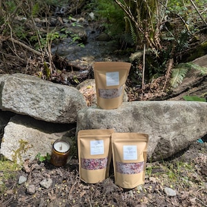 May include: Four brown paper pouches of bath salts with a white label that reads "The Grove". A lit candle in a brown glass jar is next to the pouches. The bath salts are pink and white. The background is a natural outdoor setting.