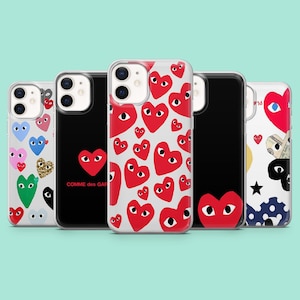 Puede incluir: Una colección de fundas para teléfonos con varios diseños. Las fundas presentan gráficos de corazones rojos con ojos, fundas negras con gráficos de corazones y fundas con patrones de corazones coloridos. El texto "COMME des GAR" es visible.