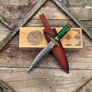 Puede incluir: Una daga artesanal con una hoja estampada y un mango verde, apoyada sobre una funda de cuero marrón. La daga se exhibe con una caja de madera con un diseño grabado. La estética general es rústica y artesanal.