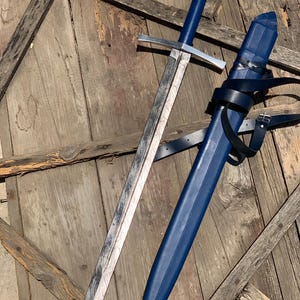 May include: A longsword with a patterned Damascus steel blade, a straight silver crossguard, and a blue leather-wrapped grip. The pommel is a silver wheel shape. A matching blue leather scabbard with a silver tip and black leather straps is included.