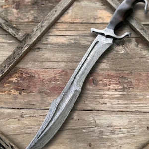 May include: A curved sword featuring a patterned Damascus steel blade with a distinct wavy texture. The hilt is crafted from dark, smooth material and includes a metal crossguard and a hooked pommel at the end of the handle for a secure grip.