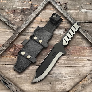 Puede incluir: Un cuchillo de hoja fija con acabado en negro y plata, que presenta un lomo dentado y un mango esqueletizado. A su lado se encuentra una funda negra con un patrón texturizado inspirado en reptiles, asegurada por cuatro botones metálicos plateados.