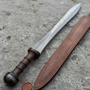 May include: A detailed image of a Roman gladius sword with a patterned steel blade and a dark wooden handle. The sword rests beside a brown leather sheath with decorative stitching. A small, round seal is attached to the handle.
