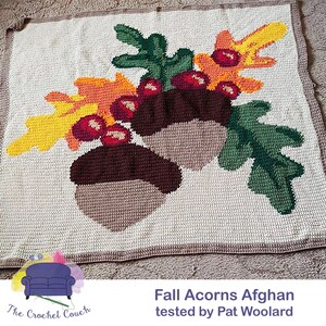 May include: Crocheted blanket featuring a fall-themed design with acorns, leaves, and berries in shades of brown, green, orange, and yellow. The blanket is made with a textured stitch and has a white background. The text "Fall Acorns Afghan tested by Pat Woolard" is visible at the bottom of the image.