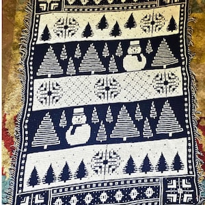May include: A rectangular crochet blanket featuring a dark blue and white color scheme with repeating horizontal bands. The design includes motifs of pine trees, snowmen, and geometric snowflakes. The text "crocheted by Tanya Watt" is visible.