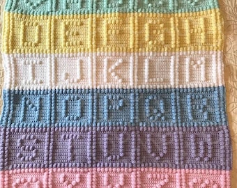 Puede incluir: Una manta rectangular de ganchillo con un diseño de rayas multicolores en verde menta, amarillo, blanco, azul, morado y rosa. Cada fila contiene letras del alfabeto y formas de corazón en relieve hechas con punto garbanzo.