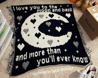 May include: A black and white crocheted blanket featuring a large crescent moon, scattered hearts, and stars. The text reads I love you to the moon and back and more than you'll ever know. The blanket has fringed edges on the left and right sides.