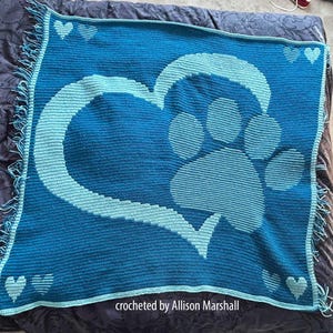 Könnte beinhalten: Eine blaue Häkeldecke mit einem Herz- und Pfotenabdruck-Design in einem helleren Blauton. Die Decke hat einen Fransenrand und kleine Herzakzente in jeder Ecke. Der Text "crocheted by Allison Marshall" ist sichtbar.