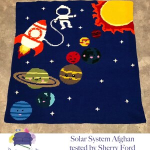 May include: A crocheted blanket depicting a solar system with a blue background, white stars, a yellow sun, a red planet, a brown planet with rings, a green planet, a blue planet, and an astronaut in a white spacesuit holding onto a red and white rocket. The text "Solar System Afghan tested by Sherry Ford" is at the bottom of the blanket.