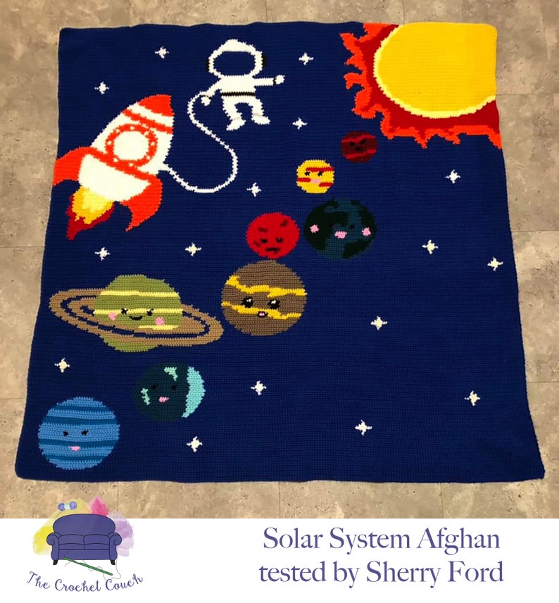 May include: A crocheted blanket depicting a solar system with a blue background, white stars, a yellow sun, a red planet, a brown planet with rings, a green planet, a blue planet, and an astronaut in a white spacesuit holding onto a red and white rocket. The text "Solar System Afghan tested by Sherry Ford" is at the bottom of the blanket.