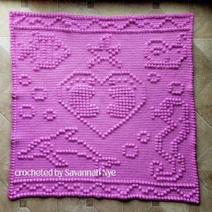 Puede incluir: Una manta cuadrada de ganchillo color púrpura con un corazón central formado por puntos de burbuja en relieve. El diseño incluye motivos marinos texturizados como un pez, una estrella de mar, una ballena y un caballito de mar, con el texto "crocheted by Savannah Nye".