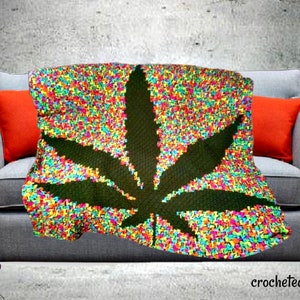 Puede incluir: Una manta de crochet colorida con un gran diseño de hoja de marihuana verde. La manta está drapeada sobre un sofá gris con almohadas naranjas. El texto "crocheted by Fawn" es visible en la esquina inferior derecha de la imagen.