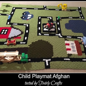 May include: A green, black, and white crocheted playmat with a road design. The playmat features a red house, a blue pond, and a white building with red stripes.  There are toy cars and trucks on the playmat. Child Playmat Afghan tested by Twirly Crafts.