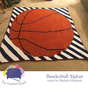 Può includere: Una coperta all'uncinetto a tema basket con strisce bianche e blu navy. La coperta presenta una grande palla da basket arancione con cuciture nere. Il testo "Basketball Afghan tested by Michele Elisabeth" è sotto la coperta.