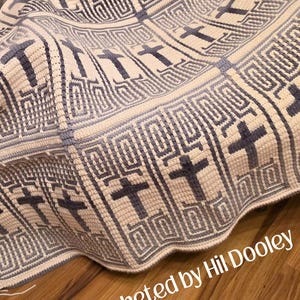 May include: A crocheted blanket with a repeating pattern of crosses in shades of gray and cream. The blanket features a textured design with geometric shapes surrounding the crosses. The text "crocheted by Hil Dooley" is visible in the bottom right corner.
