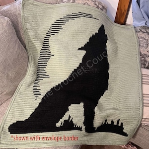 Op de afbeelding: Een groene gehaakte deken met een zwart wolfsilhouet dat naar de maan huilt. De deken heeft een zwart-wit gestreept ontwerp rond de maan. De tekst "The Crochet Couch" is zichtbaar in de afbeelding. De bijschrift "*shown with envelope border*" staat onderaan de afbeelding.