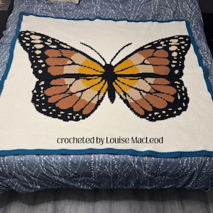 May include: A large, cream-colored crocheted blanket featuring a detailed monarch butterfly design. The butterfly has brown, black, and yellow wings. The blanket has a teal border and the text "crocheted by Louise MacLeod".