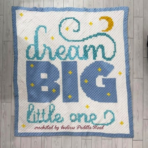 May include: A crocheted baby blanket with a blue and white background. The blanket features the words "dream big little one" in a teal and yellow color scheme. The blanket is decorated with a crescent moon and stars. The text "crocheted by Ivelisse Padilla-Head" is included at the bottom of the blanket.