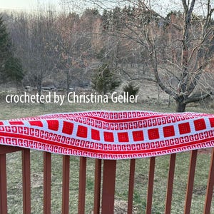 May include: A red and white crocheted scarf with a geometric pattern. The scarf is draped over a wooden railing. "Crocheted by Christina Geller" is written on the scarf.