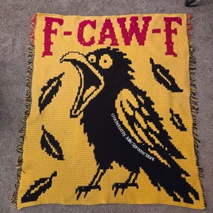 Op de afbeelding: Een rechthoekige gele gehaakte deken met een grote zwarte kraai met een open snavel. De tekst F-CAW-F is bovenaan in het rood geborduurd. Verschillende zwarte veren zijn rond de kraai verspreid. De randen hebben franjes van geel en zwart garen.