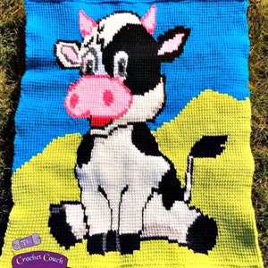 Op de afbeelding: Een vierkant gehaakt kleed met een cartoon koeienontwerp. De koe is zwart en wit met roze accenten, tegen een blauwe lucht en gele gras achtergrond. De deken heeft de tekst "The Crochet Couch" en "tested by Sherry Ford".