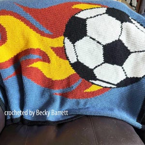 Pode incluir: Uma manta azul de crochê com um desenho de bola de futebol e chamas vermelhas, amarelas e laranjas. A manta tem uma borda preta e está sobre um sofá de couro marrom. O texto "crocheted by Becky Barrett" é visível.